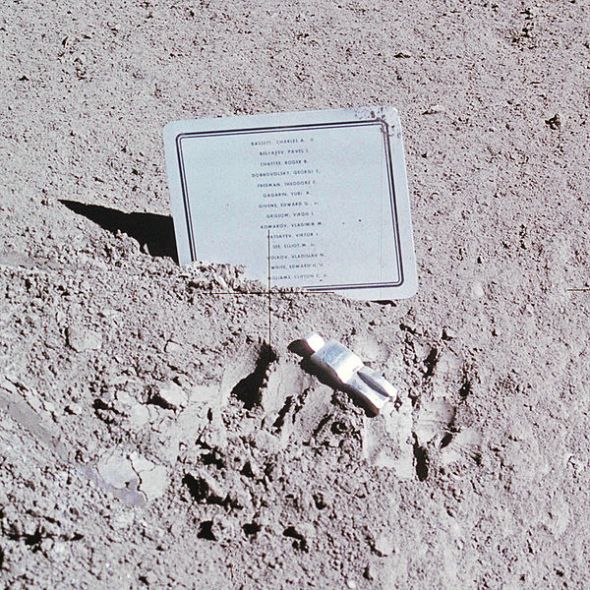Metal plaque listing names and abstract metal figure lying on the surface of the moon next to astronaut boot prints in the dust.